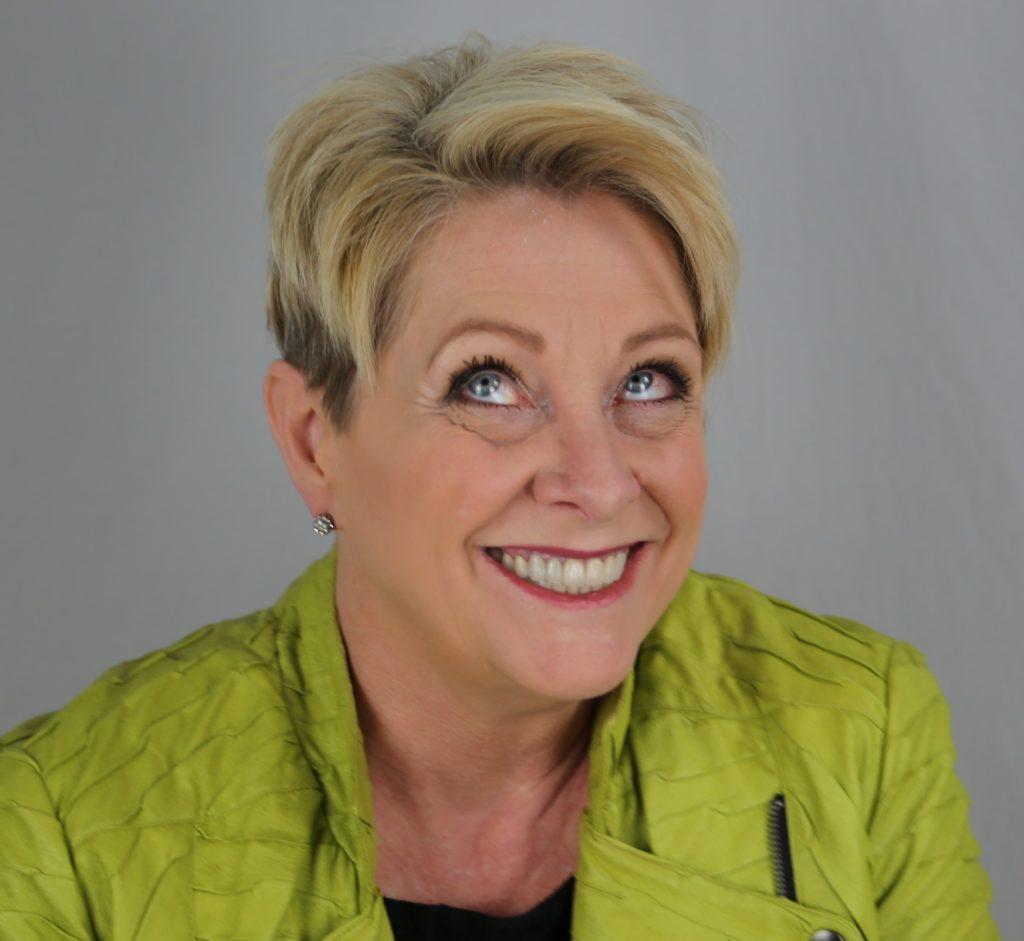 A photo of Patti Blackstaffe smiling with a goofy look in her eyes. She is wearing a green jacket brightly juxtaposed against a grey background.